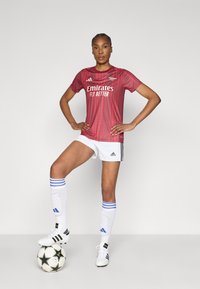 Κόκκινη φανέλα ποδοσφαίρου με λογότυπο «Emirates Fly Better», συνδυασμένη με λευκό σορτς και κάλτσες με μπλε ρίγες, ασπρόμαυρα παπούτσια ποδοσφαίρου και μια ποδοσφαιρική μπάλα με σχέδια.