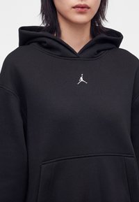 Personne portant un sweat à capuche noir avec un petit logo blanc Jumpman sur la poitrine et une poche kangourou à l'avant.