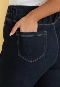 Jeans de mezclilla oscura con una cintura elástica, bolsillos laterales y costuras amarillas en contraste. El bolsillo trasero presenta un diseño rectangular clásico.