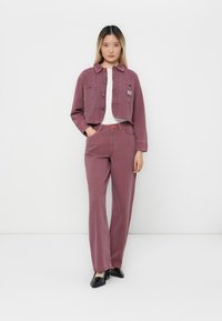 Donna che indossa un completo coordinato in velluto a coste color malva, con una maglietta bianca e scarpe nere, in piedi su uno sfondo bianco.