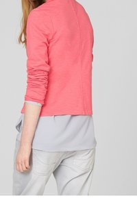 Top rosa a maniche lunghe con vestibilità morbida e scollatura rotonda, sovrapposto a una camicia grigia chiaro. Dettaglio con bottone argento sul polsino. Stile casual.