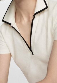 Polo blanc avec une bordure noire, doté d'un col plat et d'une courte fermeture éclair. Le tissu semble texturé et légèrement structuré.