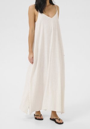 Woman wearing a sleeveless, white maxi dress with thin black vertical stripes and black flip-flop sandals, standing on a plain background.