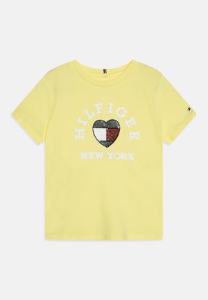 Žluté bavlněné tričko s texturovaným černým srdcem, které má červené, bílé a modré části, a bílým textem "HILFIGER NEW YORK" pod ním.