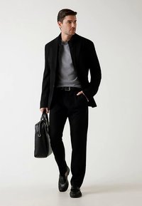 Blazer structuré noir sur un t-shirt gris à col rond, associé à un pantalon noir sur mesure. Des baskets noires et une mallette en cuir complètent le look.