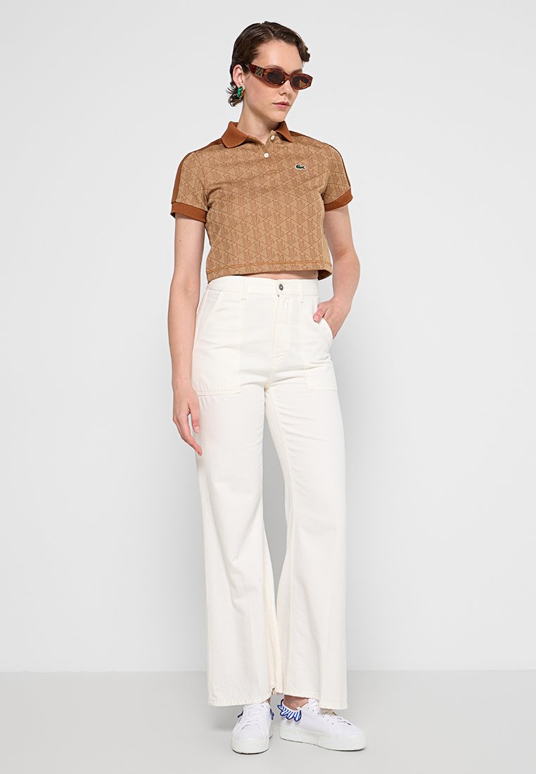 Camisa polo marrón con estampado y cuello marrón, pantalones anchos blancos y zapatillas blancas. Los accesorios incluyen gafas de sol marrones y pendientes.