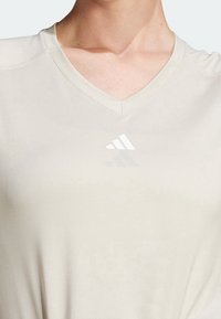 V-ringad sporttröja i ljusbeige tyg, med en subtil textur och en liten vit Adidas-logga på bröstet.