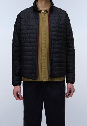 Personne portant une veste matelassée noire à fermeture éclair par-dessus une chemise olive à boutons et un pantalon noir, debout sur un fond clair.