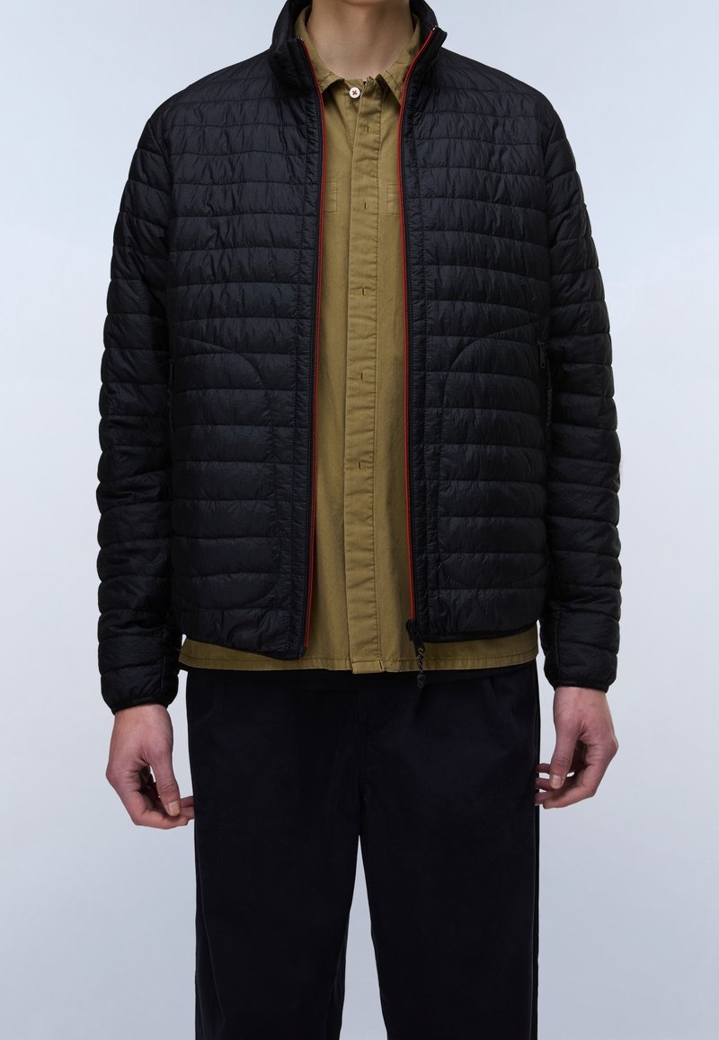 Personne portant une veste matelassée noire à fermeture éclair par-dessus une chemise olive à boutons et un pantalon noir, debout sur un fond clair.