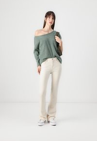 Grønn avskåret strikkegenser, kremfargede vide jeans, hvite sneakers. Modellen poserer med en veske, og viser frem et uformelt antrekk mot en ensfarget bakgrunn.