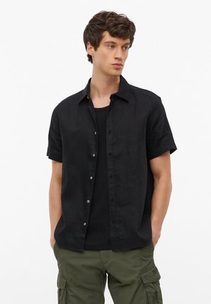 Młody mężczyzna z krótkimi kręconymi włosami, noszący rozpiętą czarną koszulę z krótkim rękawem na czarnym t-shircie i oliwkowozielone spodnie typu cargo, ręce w kieszeniach.