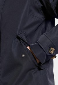 Marinblå vattentät jacka med en slät textur, med knappad manschett, sidoficka och logotypdetaljer på ärmen.