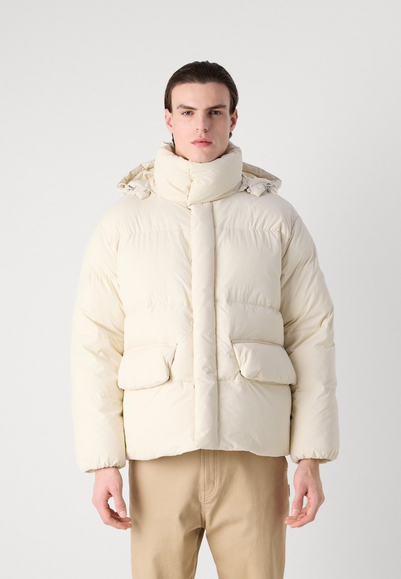 Giacca a vento in crema, con colletto alto, due tasche laterali e una silhouette ampia. Realizzata in materiale morbido e imbottito.