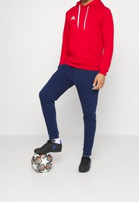 Rotes Kapuzen-Sweatshirt, marineblaue Jogginghose und schwarze Fußballschuhe, die auf einem gemusterten Fußball stehen. Enthält Adidas-Logos auf der Kleidung.