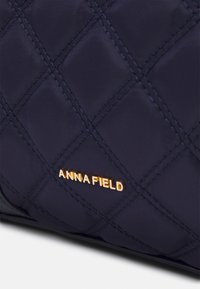 Marinblå vadderad tyghandväska med guld "ANNA FIELD"-logotyp, som visar en texturerad yta och jämnt fördelade diamantmönster.