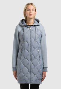 Fiatal nő világoskék steppelt hosszú kabátban, kapucnival, elöl cipzárral és cipzáras zsebekkel, egyszínű háttér előtt állva.