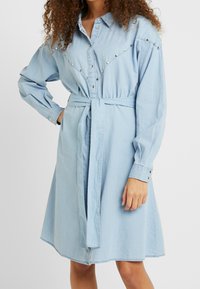 Light blue denim shirt dress with long sleeves, buttoned cuffs, a belted waist, and metal studs along the yoke and shoulders.