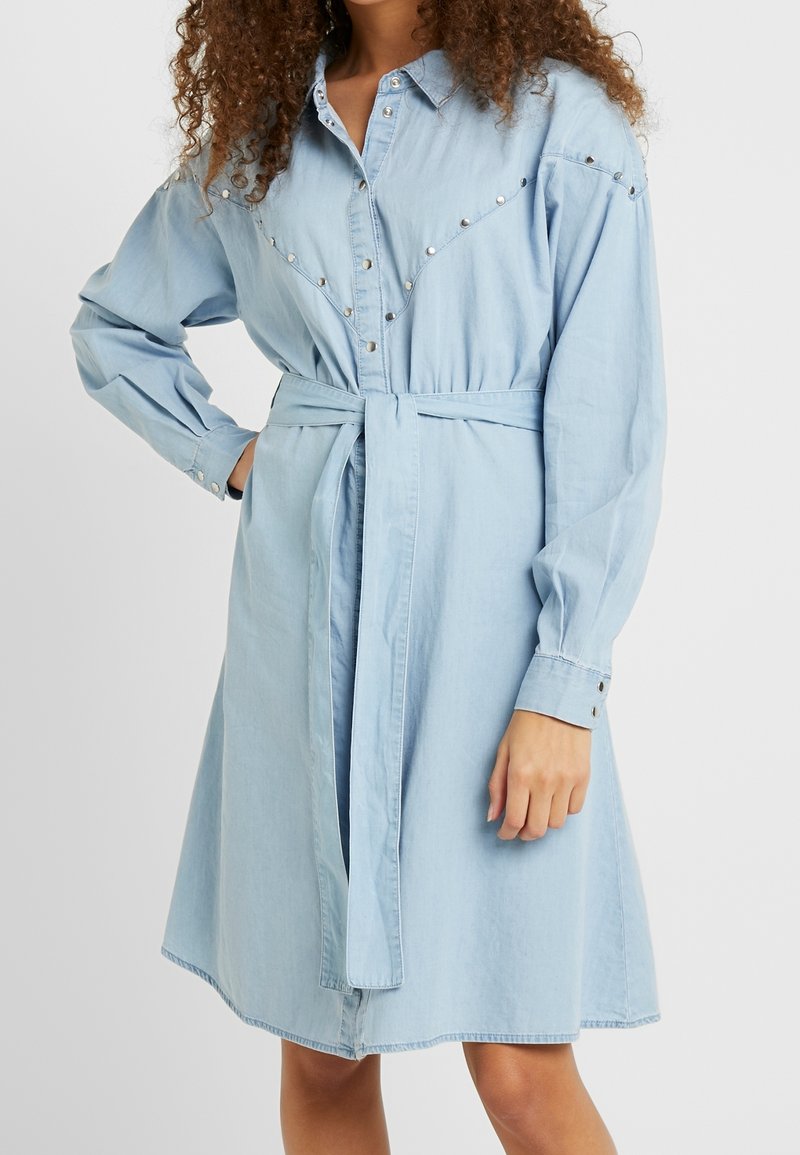 Light blue denim shirt dress with long sleeves, buttoned cuffs, a belted waist, and metal studs along the yoke and shoulders.