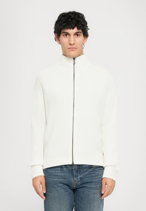 Mann iført en hvit ribbet jakke med høy krage og blå jeans, stående mot en enkel hvit bakgrunn.