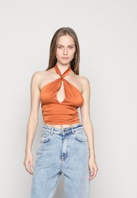 Top halter cor de laranja com um nó na frente e detalhe em forma de chave, apresentando um tecido liso e elástico. Usado com jeans de cintura alta azul claro.