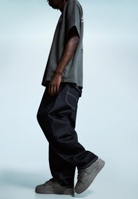 Prebujné šedé bavlnené tričko, voľné tmavozelené denimové nohavice a šedé tenisky. Opotrebované textúry, kontrastné stehy, viditeľné detaily bočných vreciek.