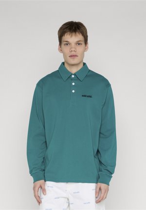 Joven con una camiseta tipo polo de manga larga color verde azulado con tres botones blancos y pantalones blancos con patrones de texto azul.