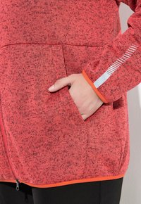 Veste rouge texturée zippée avec des contours orange contrastants. Présente une poche latérale et des bandes réfléchissantes sur la manche pour une meilleure visibilité.