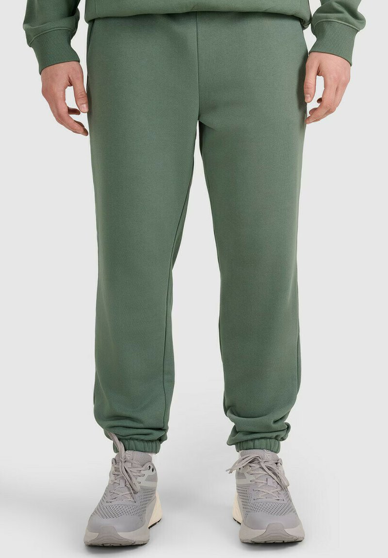 Nogi w zielonych spodniach dresowych z elastycznymi mankietami i jasnoszarych sportowych trampkach na sznurowadła na jednolitym tle.