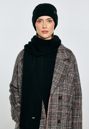 Czarna dziergana czapka i szalik o prążkowanej fakturze, zestawione ze sztucznym wełnianym płaszczem w kratę z wzorami w odcieniach szarości, czerni i czerwieni.