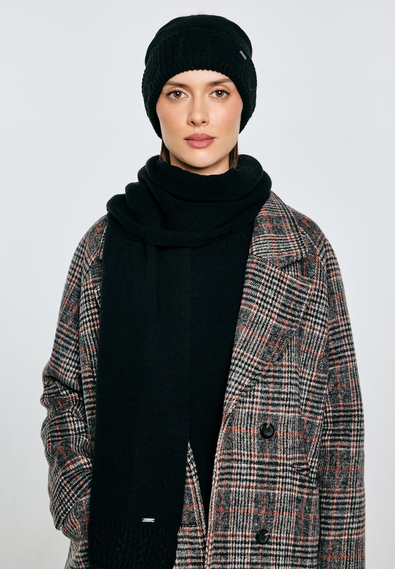 Czarna dziergana czapka i szalik o prążkowanej fakturze, zestawione ze sztucznym wełnianym płaszczem w kratę z wzorami w odcieniach szarości, czerni i czerwieni.