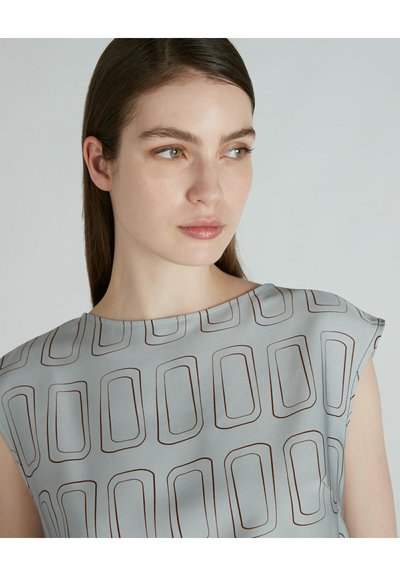 Top grigio chiaro con un sottile motivo geometrico marrone. Il tessuto appare liscio con maniche a cappuccio e un ampio scollo.