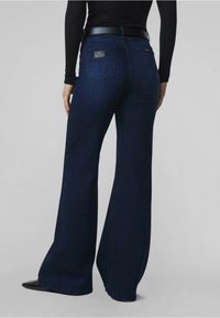 Vaqueros acampanados de color azul oscuro, hechos de mezclilla, con una superficie texturizada, dos bolsillos traseros y un discreto parche de logo cerca de la cinturilla.