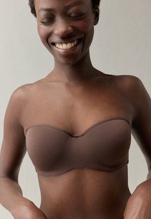 Lachende donkerhuidige vrouw met gesloten ogen, die een strapless bruine bh draagt tegen een effen lichte achtergrond.