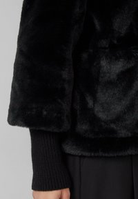 Veste en fausse fourrure noire avec une texture douce et des poignets en tricot côtelé. Le design présente une coupe décontractée et des coutures minimales, mettant en avant la simplicité.