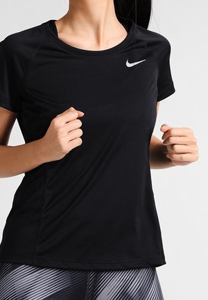Femme portant un t-shirt de sport noir à manches courtes Nike et un legging à motifs, montrée du cou aux hanches avec les mains levées comme si elle faisait du jogging.