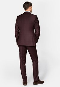 Bordowy garnitur w kratkę o dopasowanym kroju, z teksturowanej tkaniny, z wyraźnie zaakcentowanymi ramionami oraz dopasowanymi spodniami w zestawie z brązowymi butami.