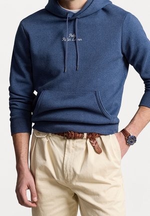 Mann trägt eine marineblaue Polo Ralph Lauren Kapuzenjacke mit Fronttasche, beige plissierte Hose, braunen geflochtenen Gürtel und eine Armbanduhr mit Lederarmband.