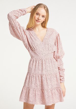 Junge Frau mit langen blonden Haaren, die ein rosafarbenes langärmliges Kleid mit kleinen schwarzen Herzmustern trägt, posiert mit einer Hand hinter dem Kopf.