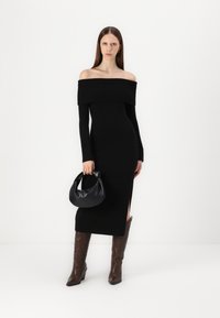 Vestido preto c/ ombros à mostra, com textura canelada, mangas longas e racha lateral. Combinado com uma mala hobo preta e botas de salto castanhas.