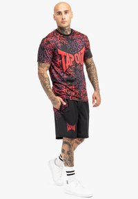 Rød og sort grafisk T-shirt med "TAPOUT" logo; korte sorte shorts; hvide sneakers; fremtrædende tatoveringer på arme og ben.