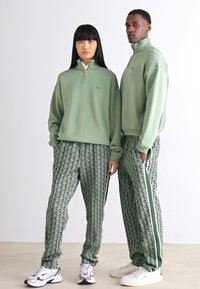 Duas modelos usam sweatshirts de zíper verde claro combinando e calças verdes estampadas com riscas brancas. O tecido parece suave e texturizado.