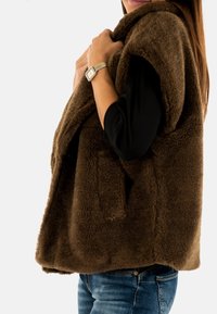 Bruine, fluffy mouwloze vest met een zachte textuur, twee voorzakken en een ronde kraag, gecombineerd met blauwe jeans.