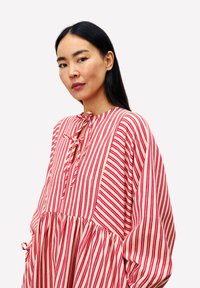 Frau mit langen schwarzen Haaren, die ein lockeres rot-weiß gestreiftes Kleid mit Schleifendetails am Ausschnitt und an den Ärmeln trägt.