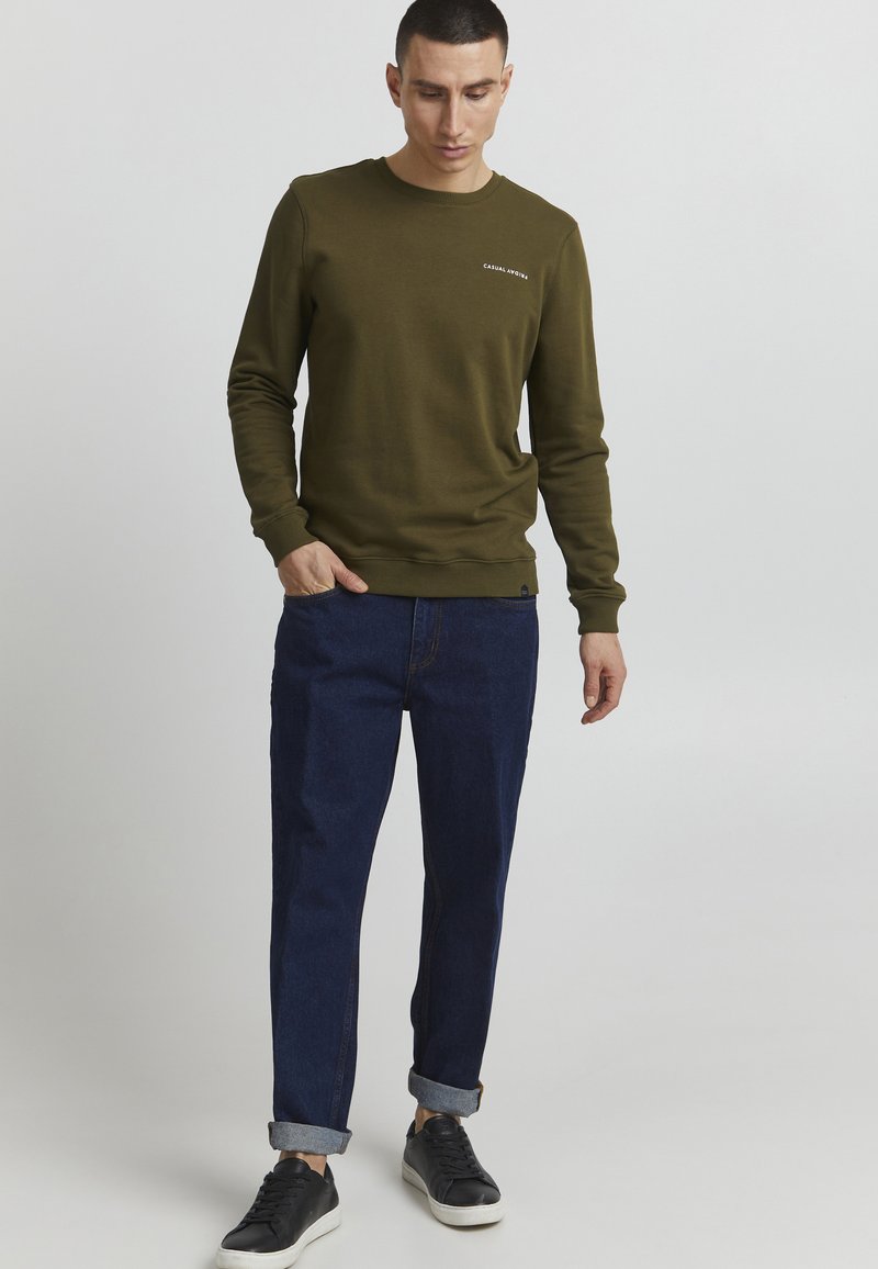 Olivenfarget sweatshirt med rund hals og strikkede mansjetter, sammen med mørkeblå oppbrettede jeans og svarte joggesko.
