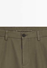 Pantalons en coton vert olive avec un devant plat, fermeture à bouton noir, passants de ceinture et poches latérales. Texture lisse et coupe ajustée.