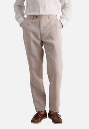 Hombre que lleva pantalones de lino beige, camisa blanca abotonada y zapatos náuticos de cuero marrón, de pie con las manos en los bolsillos.