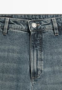 Jeans in denim blu sbiadito con chiusura a bottone, design a cinque tasche e cuciture visibili. La texture appare robusta con una leggera lucentezza.