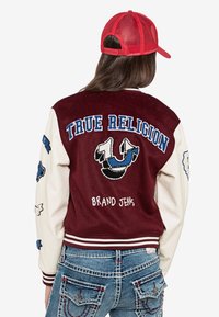 Burgundowa kurtka varsity z kremowymi rękawami, haftowanym logo "True Religion" i naszywkami. Połączona z niebieskimi dżinsami z ozdobnym przeszyciem.