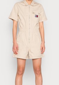 Beige krátký overal z bavlny se zipem vpředu, límečkem, elastickým pasem a dvěma bočními kapsami. Na levém prsu se nachází logo.