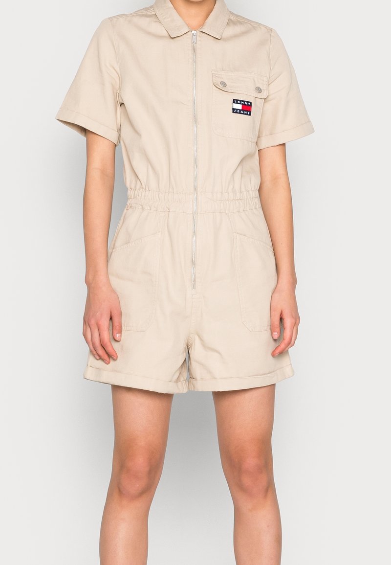 Beige krátký overal z bavlny se zipem vpředu, límečkem, elastickým pasem a dvěma bočními kapsami. Na levém prsu se nachází logo.